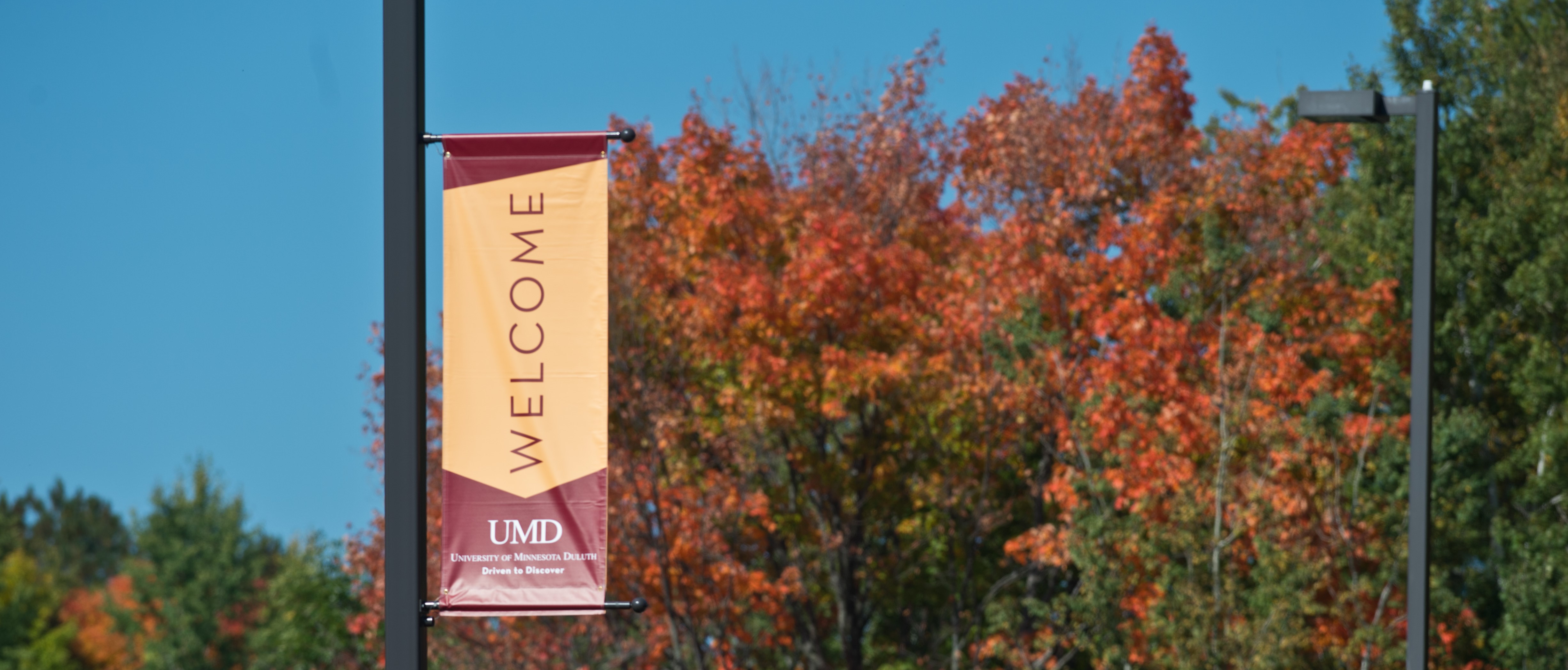 Maroon and Gold Welcome sign on a fall campus scene